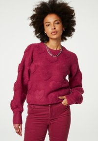 Fluffy roze trui met gestructureerde hartpatronen, geribde manchetten en zoom. Gecombineerd met bijpassende roze corduroy broek. Zilveren ketting zichtbaar.