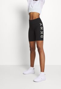 Schwarze DKNY-Logo-Shorts mit hoher Taille, aus dehnbarem Stoff, kombiniert mit weißen Sportschuhen und einem leichten Oberteil.