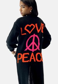 Cardigan negro de punto con grandes diseños en textil naranja y rosa que dicen "AMOR" y "PAZ", con un corazón y un símbolo de paz.