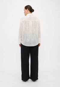 Transparent long-sleeve shirt with sequins, layered texture, white color, and stylish cuffs, paired with loose black pants.