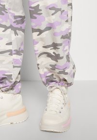 Jambes inférieures portant un pantalon camouflage beige, gris et lavande rentré dans des baskets montantes blanches cassées avec des accents beiges et roses.