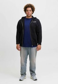 Juodas džemperis su užtrauktuku, tamsiai mėlynas marškinėlis, išblukę džinsiniai kelnės, pilki sportbačiai. Modelis stovi prieš paprastą baltą foną.