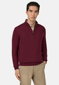 Burgunderfarbener Pullover mit Reißverschlusskragen aus weichem Stoff, mit gerippten Bündchen und Saum, kombiniert mit beigen Hosen, vor einem einfarbigen weißen Hintergrund.