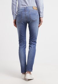 Osoba w dopasowanych niebieskich dżinsach, białych trampkach i niebiesko-białej w paski koszuli z długim rękawem, stojąca tyłem do przodu.