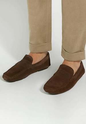 Brown suede loafers with a moccasin design, featuring a simple front band and contrast stitching, paired with tan, rolled-up trousers.