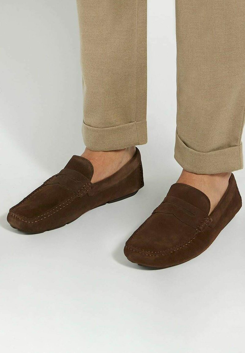 Brown suede loafers with a moccasin design, featuring a simple front band and contrast stitching, paired with tan, rolled-up trousers.