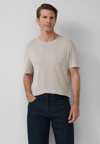 Beige T-Shirt mit kurzen Ärmeln und Rundhalsausschnitt, aus einem strukturierten Stoff. Kombiniert mit dunkelblauen Jeans, Hände in den Taschen, vor einem grauen Hintergrund.