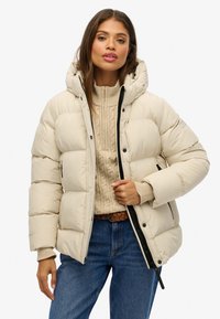 Doudoune beige avec capuche, présentant de grandes sections matelassées, une fermeture éclair frontale et deux poches zippées. Portée par-dessus un pull en tricot torsadé crème.