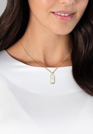Goldene Halskette mit einem rechteckigen Anhänger, der ein strahlendes Muster und einen zentralen Stein zeigt, getragen auf einem weißen Hemd.