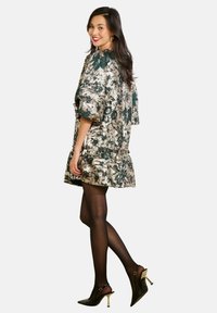 Kurzes Kleid in metallic Gold mit grünem Blumenmuster, mit Rüsche an den Ärmeln und dem Saum, kombiniert mit schwarzen Strumpfhosen und hohen Schuhen.