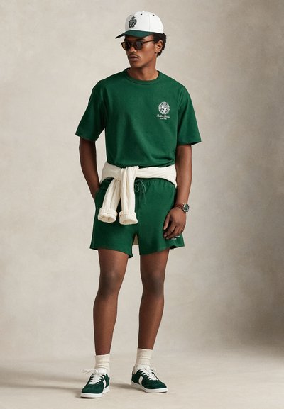 Πράσινο βαμβακερό μπλουζάκι και σορτς με λογότυπο. Κρεμ πουλόβερ δεμένο στη μέση, λευκό καπέλο και πράσινα αθλητικά παπούτσια με λευκά κορδόνια.