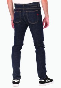 Homme portant un jean slim bleu foncé et des baskets noir et blanc, se tenant de dos sur un fond blanc.