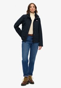 Marineblauwe waterdichte jas met een ritssluiting aan de voorkant, hoge kraag en logodetail, gedragen met lichtblauwe jeans en bruine veterschoenen.