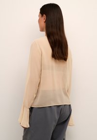 Beige läpikuultava blouse, jossa on pitkät hihnat ja kellot, väljä istuvuus ja napitettava takatuki. Valmistettu kevyestä, ilmavasta kankaasta.