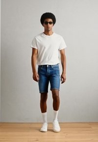 T-shirt blanche à manches courtes, shorts en denim bleu et baskets blanches. Le modèle se tient devant un fond neutre avec des lunettes de soleil et des cheveux texturés.