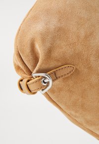 Beige semsket håndveske med en myk tekstur. Har en lærrem med sølvfarget hardware og en rynket design på den ene siden.