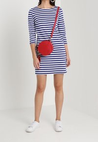 Blauw-wit gestreepte jurk met driekwartmouwen, aansluitende pasvorm, vergezeld van een ronde rode schoudertas en witte sneakers.