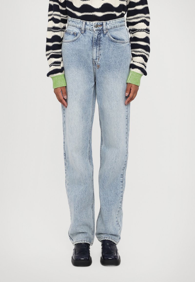 Jeans in denim azzurro chiaro con design a gamba dritta, vita alta e dettagli di cucitura visibili. Indossati con un maglione a righe e scarpe scure.