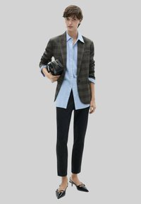 Veste à carreaux sombres sur une chemise bleu clair, assortie à un pantalon noir slim et des escarpins noirs pointus, tenant un petit sac noir.