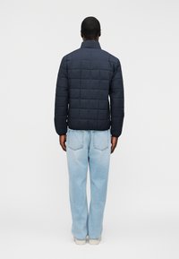 Άνδρας με κοντά μαλλιά φοράει σκούρο μπλε μπουφάν με καπιτονέ, φωτεινό μπλε τζιν παντελόνι κανονικής γραμμής και λευκά αθλητικά παπούτσια, στέκεται γυρισμένος προς τα πίσω σε λευκό φόντο.