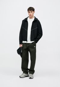 Czarna kurtka z refleksyjnymi akcentami, biała koszulka, oliwkowozielone spodnie typu cargo i szare tenisówki. Trzyma czarną czapkę. Minimalistyczny design.