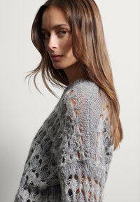 Maglione grigio chiaro lavorato a maglia con un motivo allentato e traforato e una finitura testurizzata, caratterizzato da un ampio scollo e spalle scese.