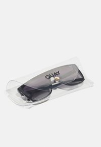Melni taisnstūra saulesbrilles, kas iekļautas caurspīdīgā plastmasas korpusā ar zelta pogas aizdari un zīmola nosaukumu "QUAY" izteikti redzamu.
