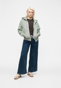 Femme blonde debout, les mains dans les poches, portant un sweat à capuche vert GAP, un t-shirt noir, un jean large bleu et des baskets beige à motifs sur un fond blanc.