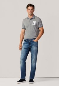 Hombre de pie con una mano en el bolsillo, vistiendo una camiseta polo gris con un parche "F", jeans azules y zapatillas negras, contra un fondo liso.