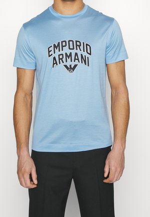 Vaaleansininen puuvillapaita, jossa on musta grafiikkaprintti "EMPORIO ARMANI" ja logo keskellä. Lyhyet hihat ja pyöreä pääntie.