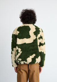 Jaqueta de fleece com padrão de camuflagem verde e creme, com uma textura suave, colarinho arredondado e mangas longas.
