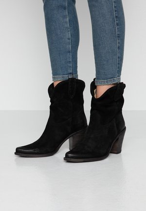 Botas de tobillo de ante negro con una parte superior curvada, punta afilada y tacón de madera apilada. Presentan costuras decorativas y lengüetas laterales.