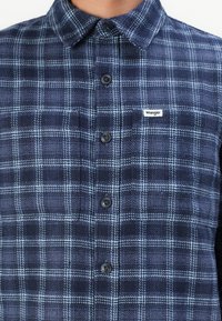 Camisa de franela de cuadros en azul marino y azul claro, con un cierre de botones en la parte delantera, cuello y una pequeña etiqueta "Wrangler" en el lado izquierdo del pecho.