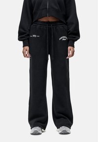 Zwarte katoenen sweatpants met een losse fit, voorzien van een elastische tailleband met trekkoord, en witte bedrukte tekst op de linker heup en rechter dij.