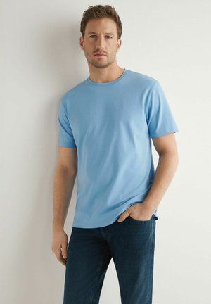 Mężczyzna w jasnoniebieskiej koszulce z krótkim rękawem i ciemnoniebieskich dżinsach, stojący na tle białej ściany z jedną ręką w kieszeni.