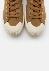 Brązowe kanvasowe trampki z kremowymi gumowymi podeszwami, okrągłym noskiem i sznurowanym wykończeniem. Posiadają metalowe oczka i teksturowaną powierzchnię materiału.