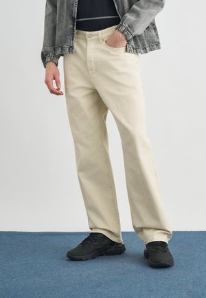 Isik, kes kannab kreemikaid sirgete jalgade pükse, musti tossusid ja halli jakki, üks käsi taskus, sinisel vaibal.