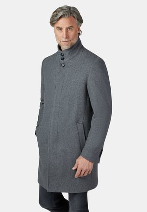 Mann trägt einen grauen Wollmantel mittlerer Länge mit hochgeknöpftem Kragen und Seitentaschen, steht mit entspannten Händen.