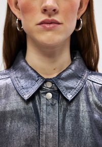 Jaqueta de ganga em azul escuro, com um colarinho estruturado, botões de metal prateados e uma textura suave com um leve brilho.