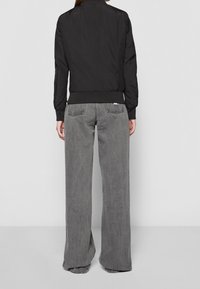 Giubbotto bomber nero con colletto e polsini a costine, abbinato a jeans in denim grigio sbiadito a gamba larga. Texture del tessuto morbido, vestibilità casual.