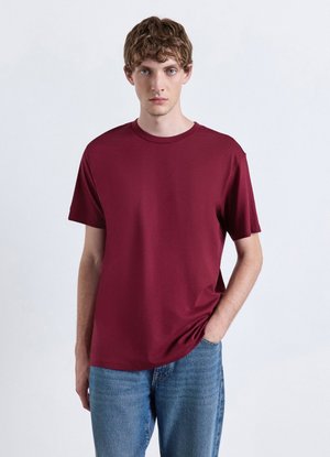 Giovane uomo con capelli ricci castani che indossa una maglietta bordeaux semplice a maniche corte e jeans blu, in piedi davanti a uno sfondo chiaro.