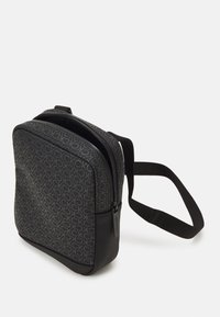 Bolso bandolera negro con patrón de logotipos repetidos, cierre de cremallera, tejido resistente y correa ajustable. Contiene una base negra texturizada.