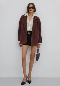 Hnědý oversized blazer, bílá pruhovaná košile, černé šortky, špičaté černé lodičky a malá černá kabelka. Hladká látka se strukturovaným designem.