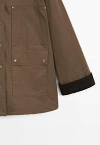 Braune Außenjacke mit zwei großen Fronttaschen, Druckknöpfen und schwarzen Cordura-Bündchen. Hergestellt aus einem strapazierfähigen, glatten Stoff.