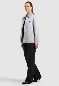 Jeune femme aux cheveux attachés portant une veste gris clair sur un haut noir, un jean noir et des chaussures noires pointues, debout de profil.