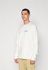Long-sleeve cream cotton t-shirt featuring a colorful "Patagonia" logo on the chest. The design is relaxed-fit with ribbed cuffs.