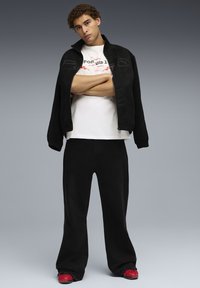 Sort zip-jakke over en hvid t-shirt med rød skrift, parret med løse sorte bukser og røde sneakers. Simpelt, afslappet look.