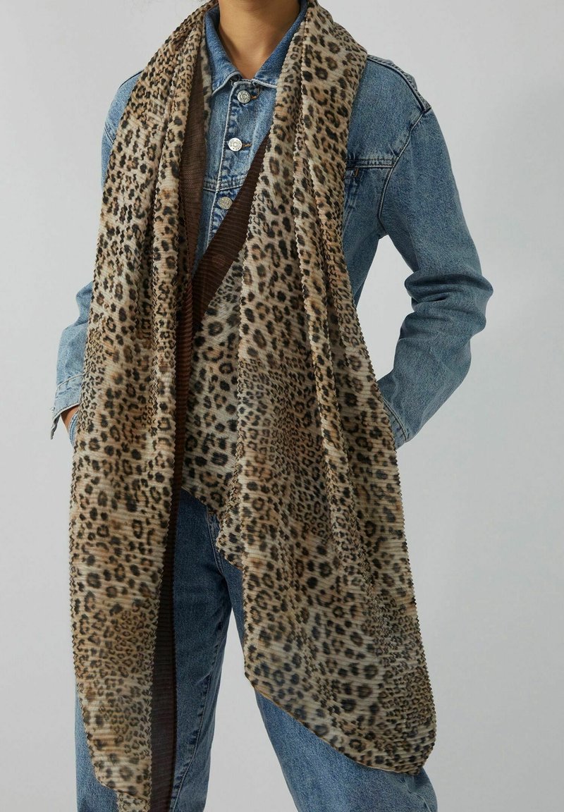 Personne portant une veste en jean bleue et un jean avec une grande écharpe à imprimé léopard drapée autour du cou et tombant sur le devant.