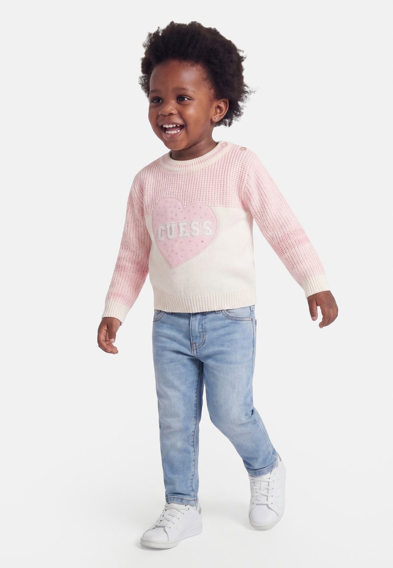Jeune enfant marchant, portant un pull rose et blanc avec un cœur et le texte "Guess", un jean bleu clair et des baskets blanches, souriant.