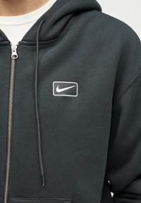 Άτομο που φοράει σκουρόχρωμο γκρι φούτερ με φερμουάρ και λευκό λογότυπο Nike στο στήθος, καθώς και λευκό T-shirt από κάτω.